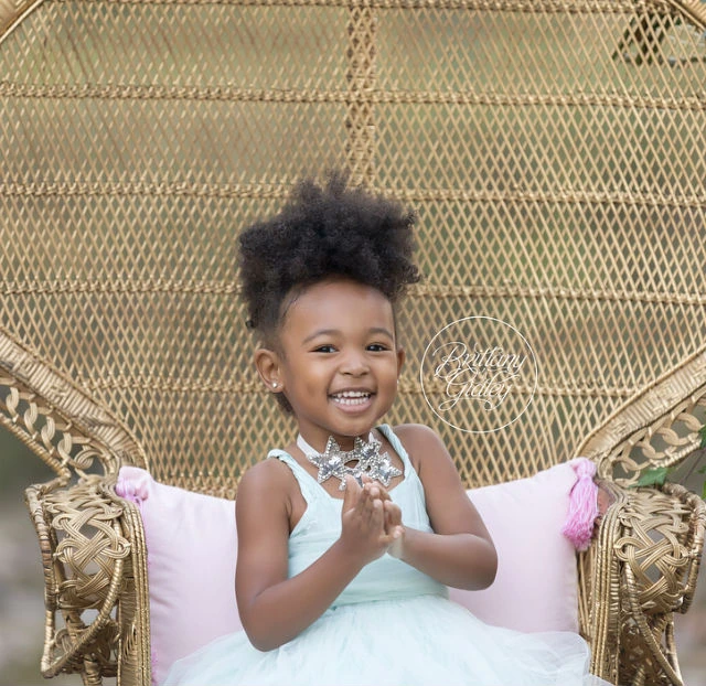 A young girl in a light blue dress smiling while seated on a wicker chair against a gold textured background.